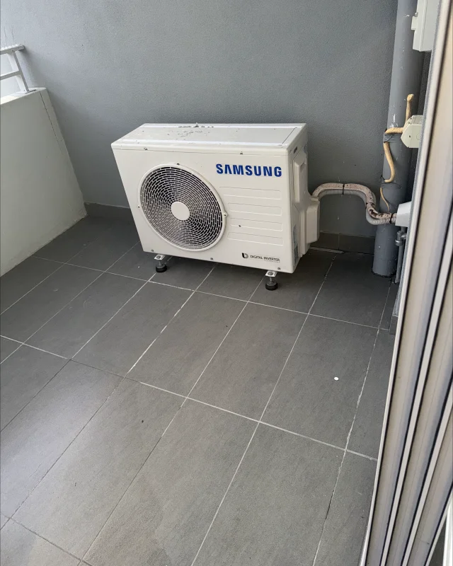 The same condenser and balcony tiles washed down clean, the unit standing in the same corner