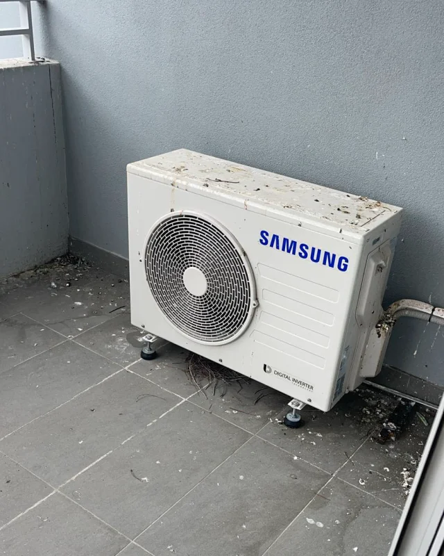 Balcony air conditioning condenser caked in bird droppings and nest debris, with grime and leaf litter across the tiled floor around it