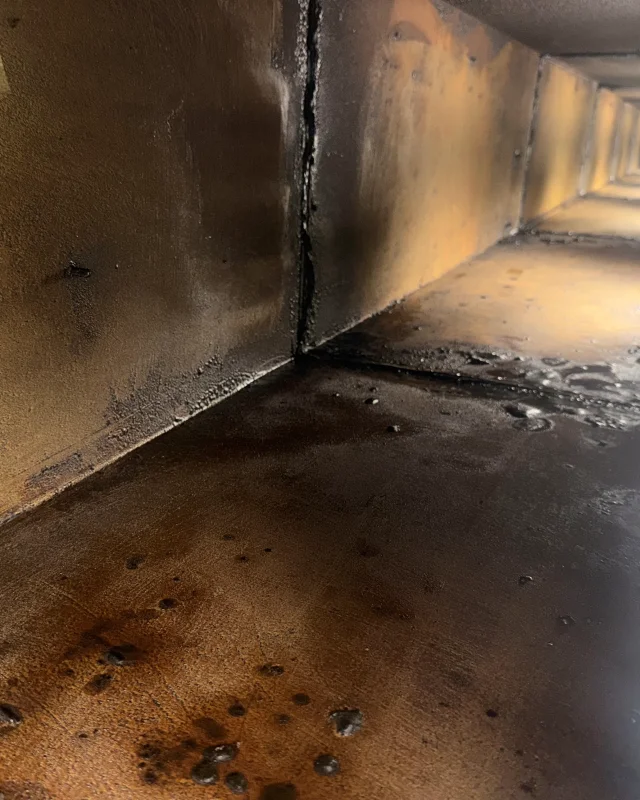 Inside a commercial kitchen exhaust duct before cleaning, hardened grease coating the walls and pooled black deposit along the base of the run
